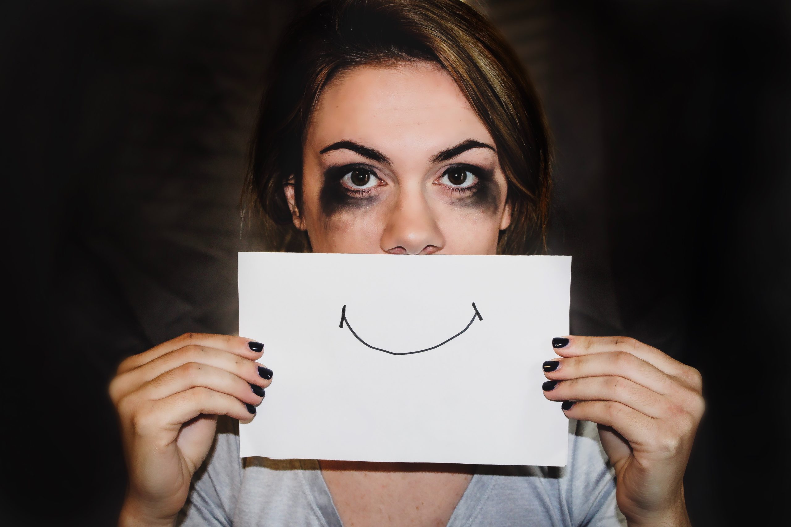 Woman with eye makeup smudged from crying holding up a smiley face paper over her mouth
